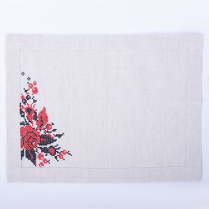 May include: A rectangular, beige linen placemat featuring a cross-stitch floral design in the upper left corner. The flowers are red and black, with green leaves. The placemat has a stitched border.