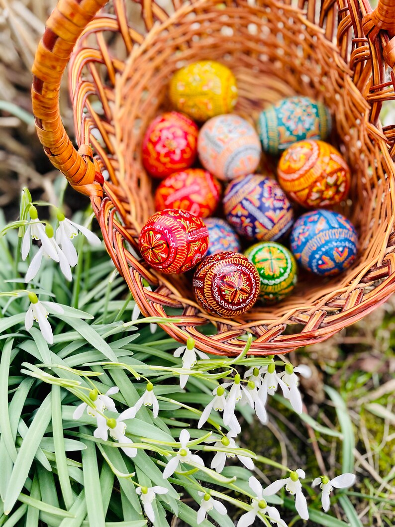 K&ouml;nnte beinhalten: Ein geflochtener Korb gef&uuml;llt mit bunt dekorierten Ostereiern. Die Eier weisen komplizierte Muster in Rot, Gelb, Blau, Orange und Gr&uuml;n auf. Wei&szlig;e Schneegl&ouml;ckchen und gr&uuml;nes Laub umgeben den Korb und schaffen eine Fr&uuml;hlingsatmosph&auml;re.