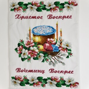 K&ouml;nnte beinhalten: Bestickter wei&szlig;er Stoff mit floralen und religi&ouml;sen Motiven. Das Design zeigt einen goldbraunen Kuchen mit blauen Streuseln, umgeben von Blumen, Kerzen und Ostereiern. Der Text "Christus ist auferstanden" und "Wahrlich, er ist auferstanden" ist rot gestickt.