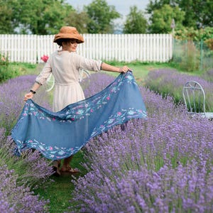 Pode incluir: Uma mulher vestindo um vestido branco e um chap&eacute;u marrom est&aacute; em p&eacute; em um campo de lavanda. Ela est&aacute; segurando um len&ccedil;o azul com um padr&atilde;o floral.