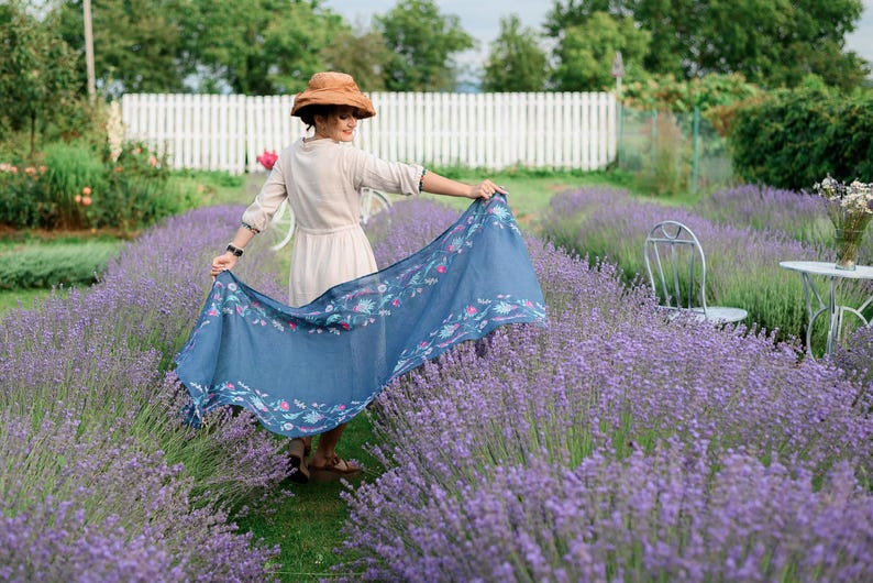 Pode incluir: Uma mulher vestindo um vestido branco e um chap&eacute;u marrom est&aacute; em p&eacute; em um campo de lavanda. Ela est&aacute; segurando um len&ccedil;o azul com um padr&atilde;o floral.