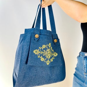 Op de afbeelding: Een blauwe denim tote bag met een geel bloemenontwerp en houten knoopsluitingen.