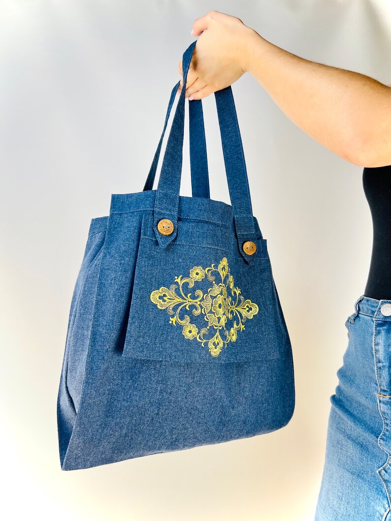 Op de afbeelding: Een blauwe denim tote bag met een geel bloemenontwerp en houten knoopsluitingen.