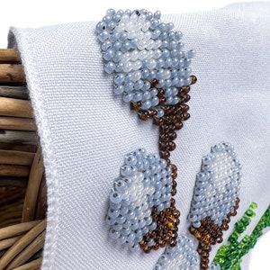 May include: Close-up of a white fabric with beaded floral embroidery. The embroidery features light blue and white beaded flowers with brown stems, and green and pink accents. The fabric is attached to a woven basket.