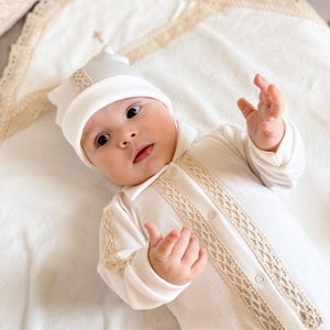 Op de afbeelding: Een witte baby romper met beige kantversiering en een witte gehaakte muts. De romper heeft een knoopsluiting en de muts heeft een klein geborduurd kruis op de bovenkant.