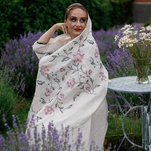 May include: A woman wearing a white shawl with floral embroidery stands in a field of lavender. The shawl has a delicate floral pattern in shades of pink and gray. The woman is looking off to the side, and her hair is pulled back from her face.