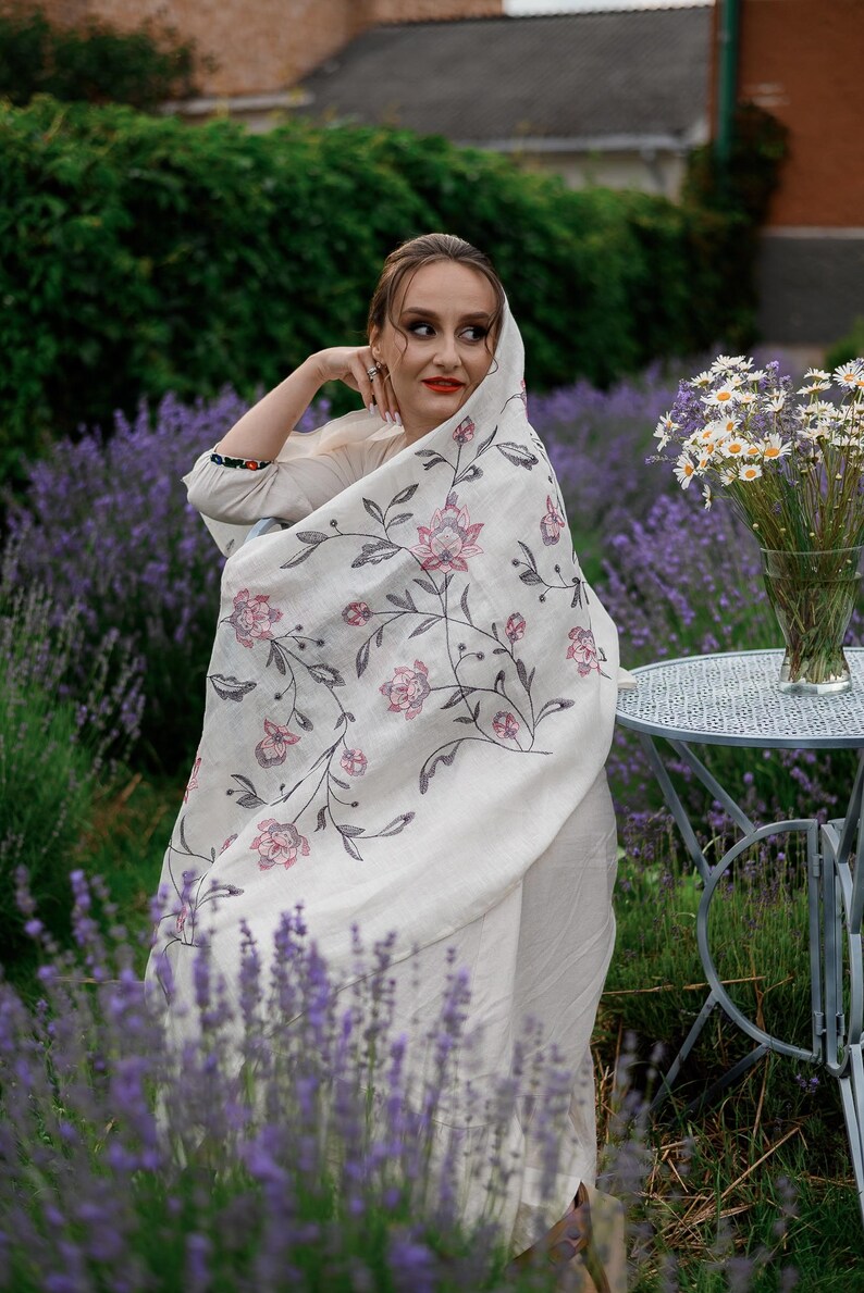 May include: A woman wearing a white shawl with floral embroidery stands in a field of lavender. The shawl has a delicate floral pattern in shades of pink and gray. The woman is looking off to the side, and her hair is pulled back from her face.