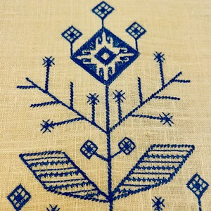 K&ouml;nnte beinhalten: Ein blaues Stickmuster auf einem beigefarbenen Leinenstoff. Das Muster zeigt eine stilisierte Blume mit einem zentralen Quadrat und vier Bl&auml;ttern.
