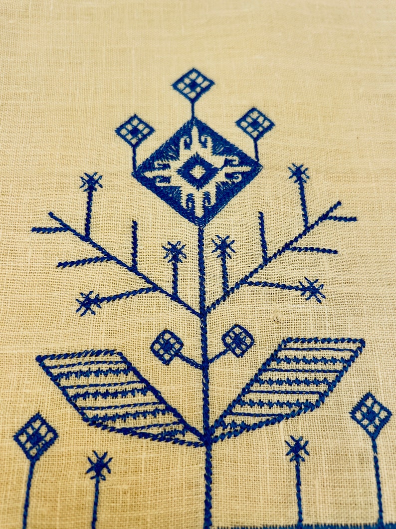 K&ouml;nnte beinhalten: Ein blaues Stickmuster auf einem beigefarbenen Leinenstoff. Das Muster zeigt eine stilisierte Blume mit einem zentralen Quadrat und vier Bl&auml;ttern.
