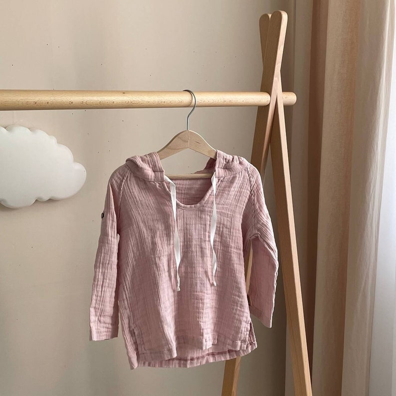May include: A pale pink hooded sweatshirt with long sleeves hanging on a wooden rack. The sweatshirt has a drawstring hood and a relaxed fit.