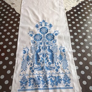 May include: White linen table runner with intricate blue floral and bird embroidery. The design features stylized flowers, leaves, and birds, with a delicate crocheted blue border along the bottom edge. A decorative piece for a dining table.
