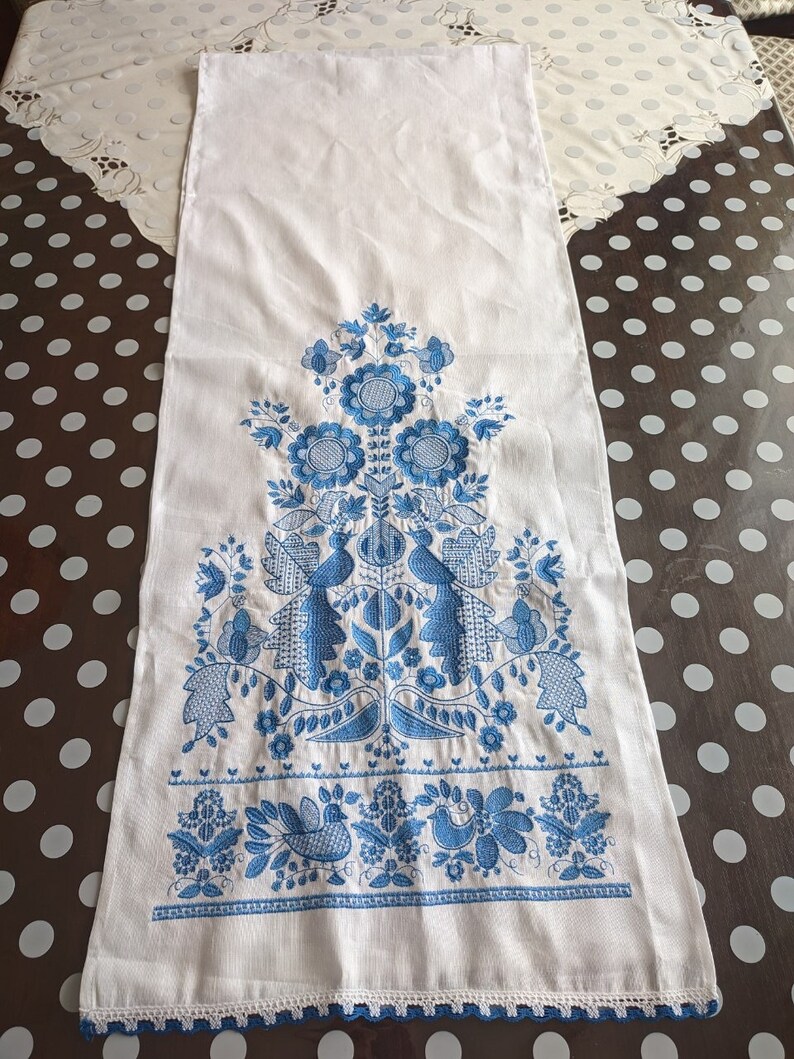 May include: White linen table runner with intricate blue floral and bird embroidery. The design features stylized flowers, leaves, and birds, with a delicate crocheted blue border along the bottom edge. A decorative piece for a dining table.