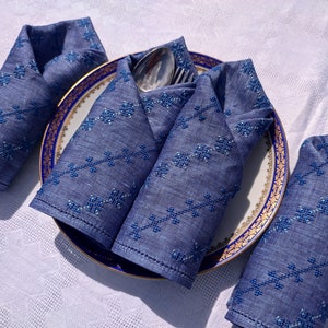 May include: A set of four folded blue linen napkins with embroidered floral patterns, arranged around a plate with a gold and blue rim. A silver fork and spoon rest on the plate, set on a white tablecloth.