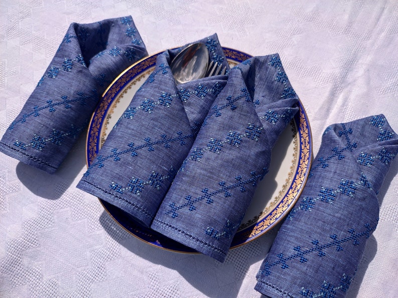 May include: A set of four folded blue linen napkins with embroidered floral patterns, arranged around a plate with a gold and blue rim. A silver fork and spoon rest on the plate, set on a white tablecloth.