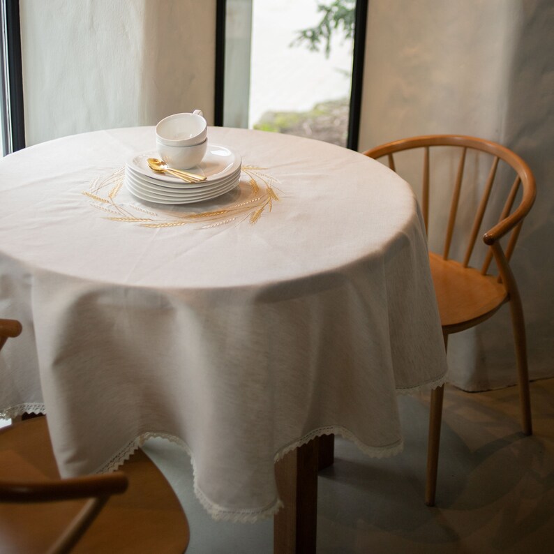 Op de afbeelding: Een ronde witte tafelkleed met een geschulpte kanten rand, op een houten tafel. Een stapel witte borden, een kopje en een gouden lepel zijn op de tafel geplaatst. Het tafelkleed heeft geborduurde tarwearen.