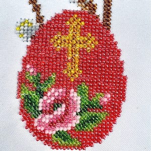 Könnte beinhalten: Ein mit Perlen besetztes Osterei-Design mit rotem Hintergrund, einem gelben Kreuz und einem rosa-grünen Blumenmuster. Das Ei ist mit kleinen, farbigen Perlen verziert und vor einem weißen Hintergrund platziert.