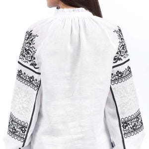 Puede incluir: Blusa blanca de manga larga con intrincados bordados florales negros en las mangas y hombros. La blusa presenta un cuello alto con volantes y un corte holgado. Las mangas tienen un dise&ntilde;o calado detallado.
