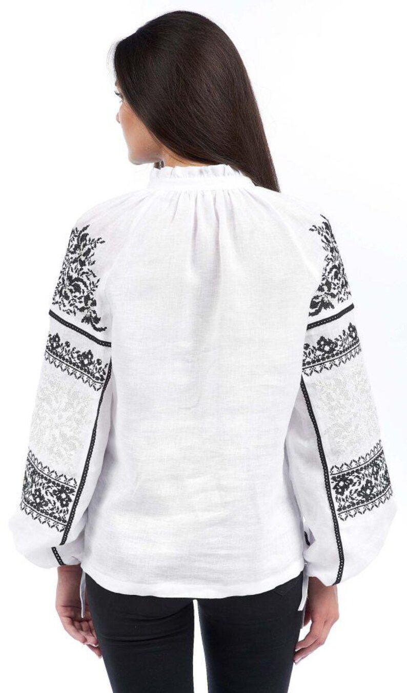 Puede incluir: Blusa blanca de manga larga con intrincados bordados florales negros en las mangas y hombros. La blusa presenta un cuello alto con volantes y un corte holgado. Las mangas tienen un dise&ntilde;o calado detallado.