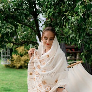 May include: A woman wearing a cream-colored shawl with a floral and striped pattern. The shawl is draped over her head and shoulders. She is wearing a cream-colored dress and has a black watch on her wrist. The background is a green yard with trees.