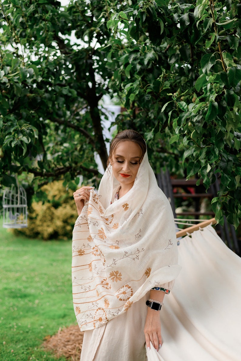 May include: A woman wearing a cream-colored shawl with a floral and striped pattern. The shawl is draped over her head and shoulders. She is wearing a cream-colored dress and has a black watch on her wrist. The background is a green yard with trees.