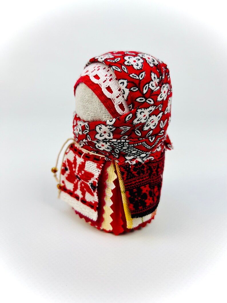 Op de afbeelding: Een traditionele Russische volkspop, bekend als een nestpop of matrjosjka, is gemaakt van stof en versierd met rode en witte bloemenpatronen. De pop is gewikkeld in een rode en witte gebloemde stoffen hoofddoek.