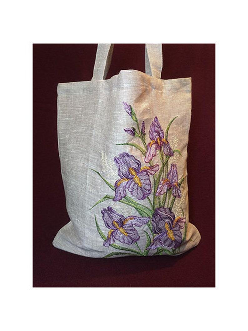 May include: A natural linen tote bag with a floral embroidery design featuring purple irises. The bag has a simple design with two handles.