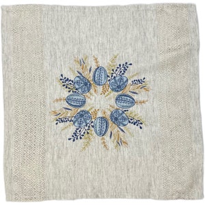 Puede incluir: Camino de mesa de lino beige con tem&aacute;tica de Pascua. El centro presenta una corona de huevos azules estampados, rodeada de follaje bordado en azul y dorado. Los bordes tienen un patr&oacute;n de diamantes.