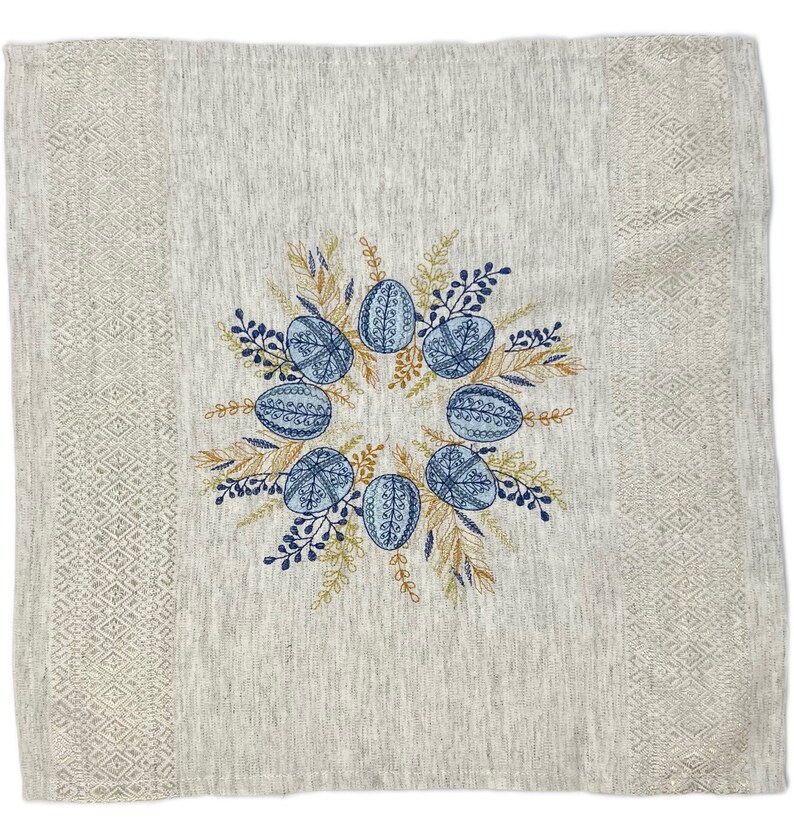 Puede incluir: Camino de mesa de lino beige con tem&aacute;tica de Pascua. El centro presenta una corona de huevos azules estampados, rodeada de follaje bordado en azul y dorado. Los bordes tienen un patr&oacute;n de diamantes.