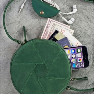 Op de afbeelding: Een ronde schoudertas van groen leer met een ritssluiting. De tas heeft een geometrisch patroon en is open om een telefoon, contant geld en kaarten te laten zien.