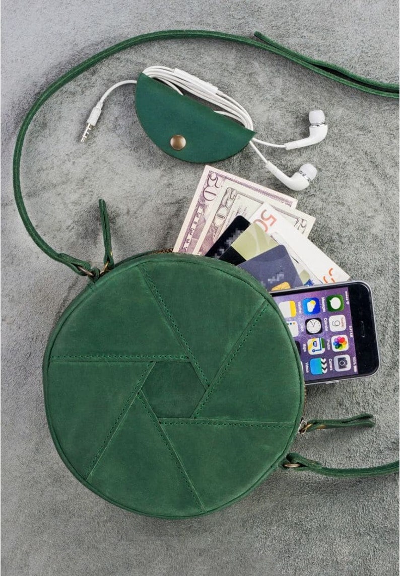 Op de afbeelding: Een ronde schoudertas van groen leer met een ritssluiting. De tas heeft een geometrisch patroon en is open om een telefoon, contant geld en kaarten te laten zien.