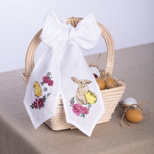 May include: White linen Easter decoration with embroidered chick and bunny designs. The chick is yellow, and the bunny is tan. Both are surrounded by pink flowers and sit on a white background. A white bow is attached to a wicker basket.