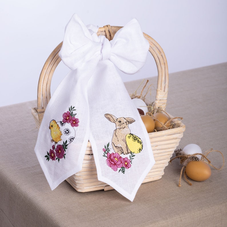 May include: White linen Easter decoration with embroidered chick and bunny designs. The chick is yellow, and the bunny is tan. Both are surrounded by pink flowers and sit on a white background. A white bow is attached to a wicker basket.