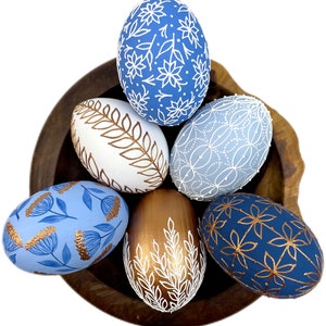 May include: Six decorated Easter eggs in a wooden bowl. The eggs are painted in shades of blue and gold with white floral and leaf designs.