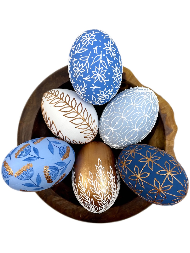 May include: Six decorated Easter eggs in a wooden bowl. The eggs are painted in shades of blue and gold with white floral and leaf designs.