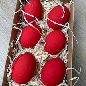 May include: A cardboard box filled with six bright red eggs nestled in white paper straw. The eggs are smooth and evenly colored, suggesting a decorative or craft item, possibly for Easter or other celebrations.