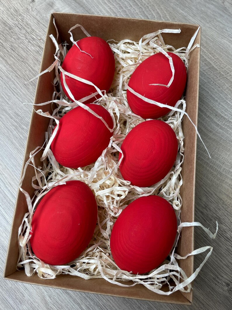 May include: A cardboard box filled with six bright red eggs nestled in white paper straw. The eggs are smooth and evenly colored, suggesting a decorative or craft item, possibly for Easter or other celebrations.