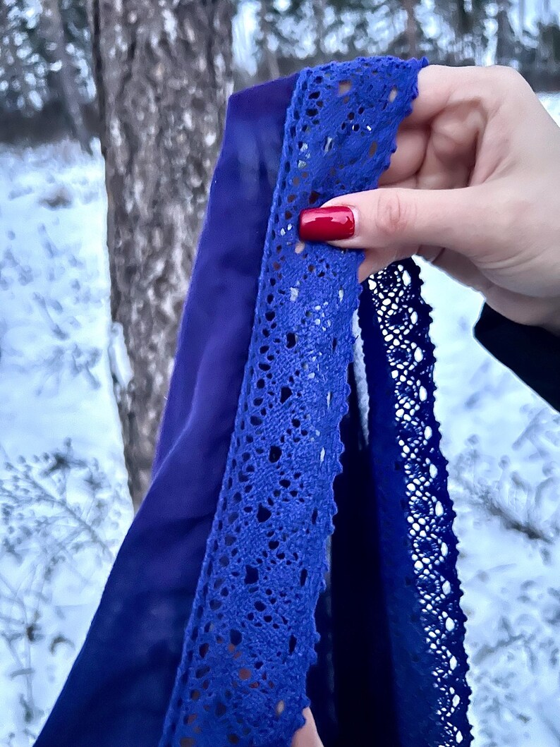 May include: A close-up of a blue fabric with a lace trim. The fabric is held in a hand with red nail polish.