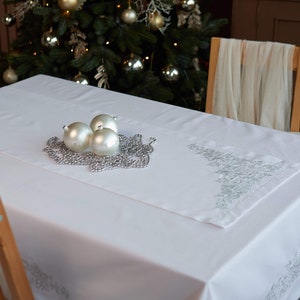 May include: A white tablecloth with a decorative silver embroidered runner. Three silver ornaments and a silver beaded garland are arranged on the table, with a Christmas tree in the background. The table is set for a holiday celebration.