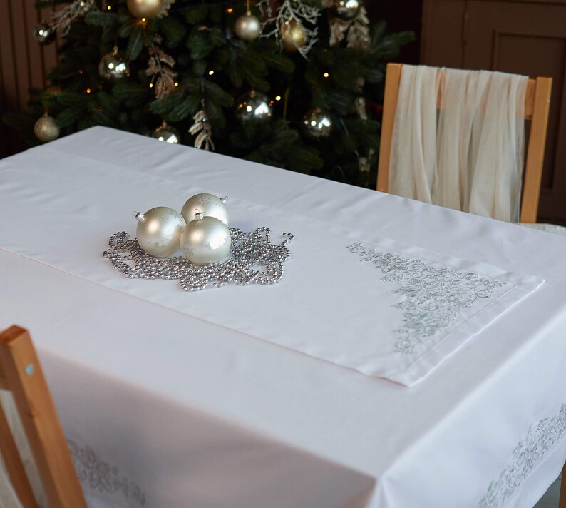 May include: A white tablecloth with a decorative silver embroidered runner. Three silver ornaments and a silver beaded garland are arranged on the table, with a Christmas tree in the background. The table is set for a holiday celebration.