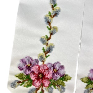 K&ouml;nnte beinhalten: Wei&szlig;es Satinband mit einem perlenbesetzten Blumendesign. Das Design umfasst rosa und violette Blumen mit gr&uuml;nen Bl&auml;ttern und einen Zweig mit hellblauen und gelben Knospen.