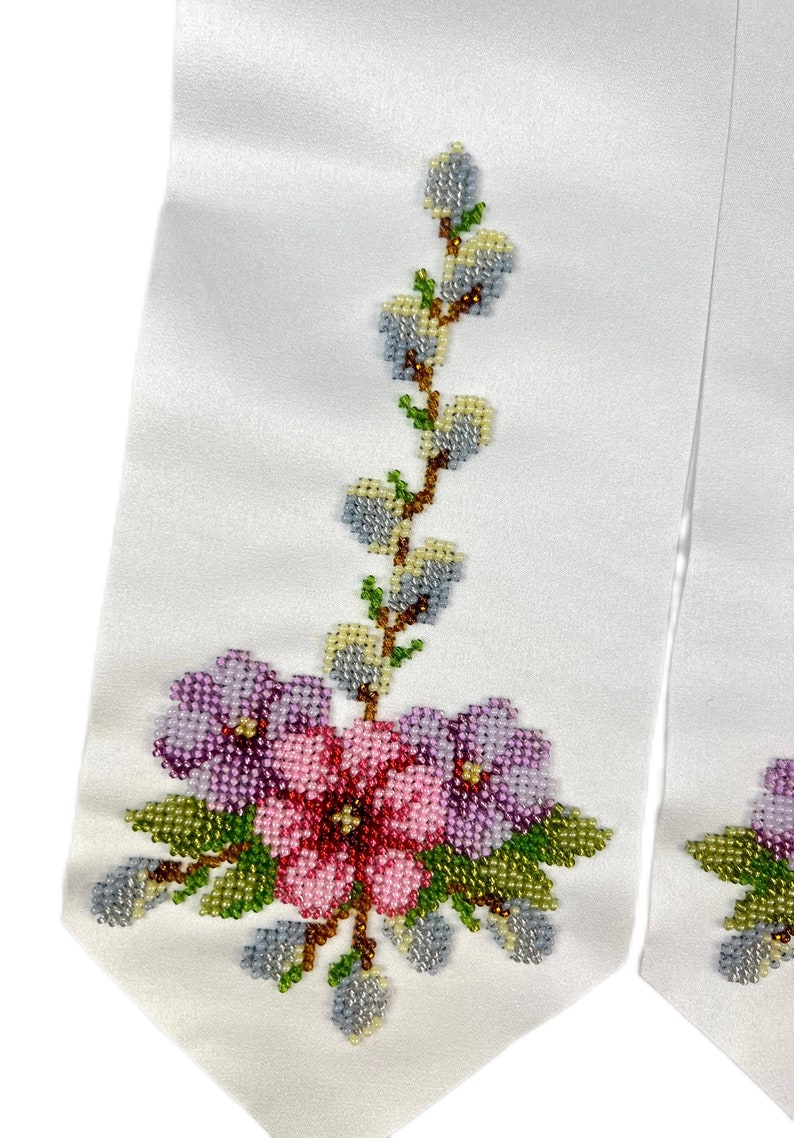 K&ouml;nnte beinhalten: Wei&szlig;es Satinband mit einem perlenbesetzten Blumendesign. Das Design umfasst rosa und violette Blumen mit gr&uuml;nen Bl&auml;ttern und einen Zweig mit hellblauen und gelben Knospen.