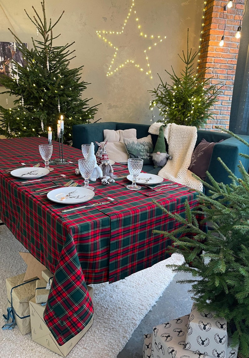May include: A festive Christmas table setting with a red and green plaid tablecloth. The table is set with plates, silverware, and crystal glasses. Decorative elements include candles, gnomes, and small Christmas trees. Presents are wrapped and placed under the table.