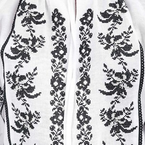 Puede incluir: Blusa blanca con bordado floral negro. La blusa presenta un cuello alto con volantes y un cierre de lazo en el escote. El bordado negro es detallado y cubre la parte delantera de la blusa.