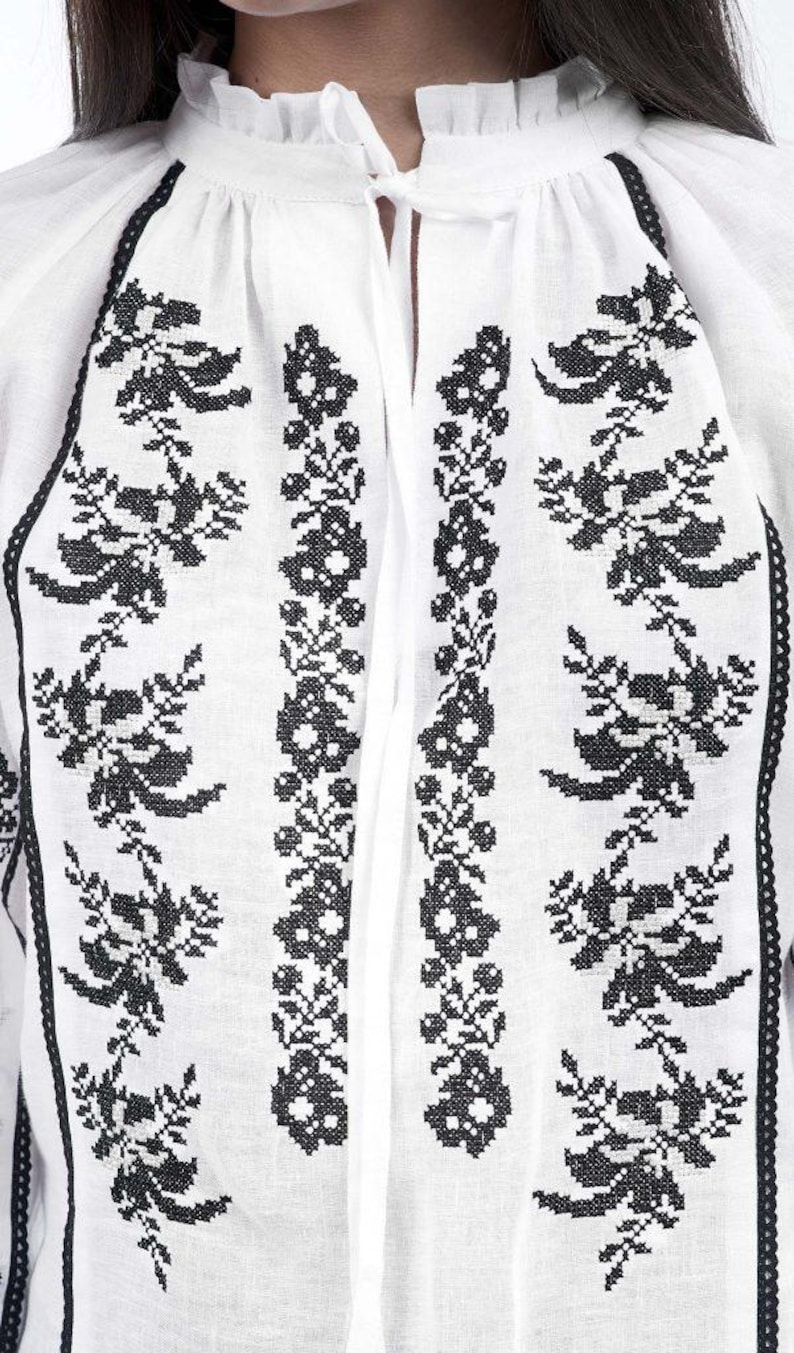 Puede incluir: Blusa blanca con bordado floral negro. La blusa presenta un cuello alto con volantes y un cierre de lazo en el escote. El bordado negro es detallado y cubre la parte delantera de la blusa.
