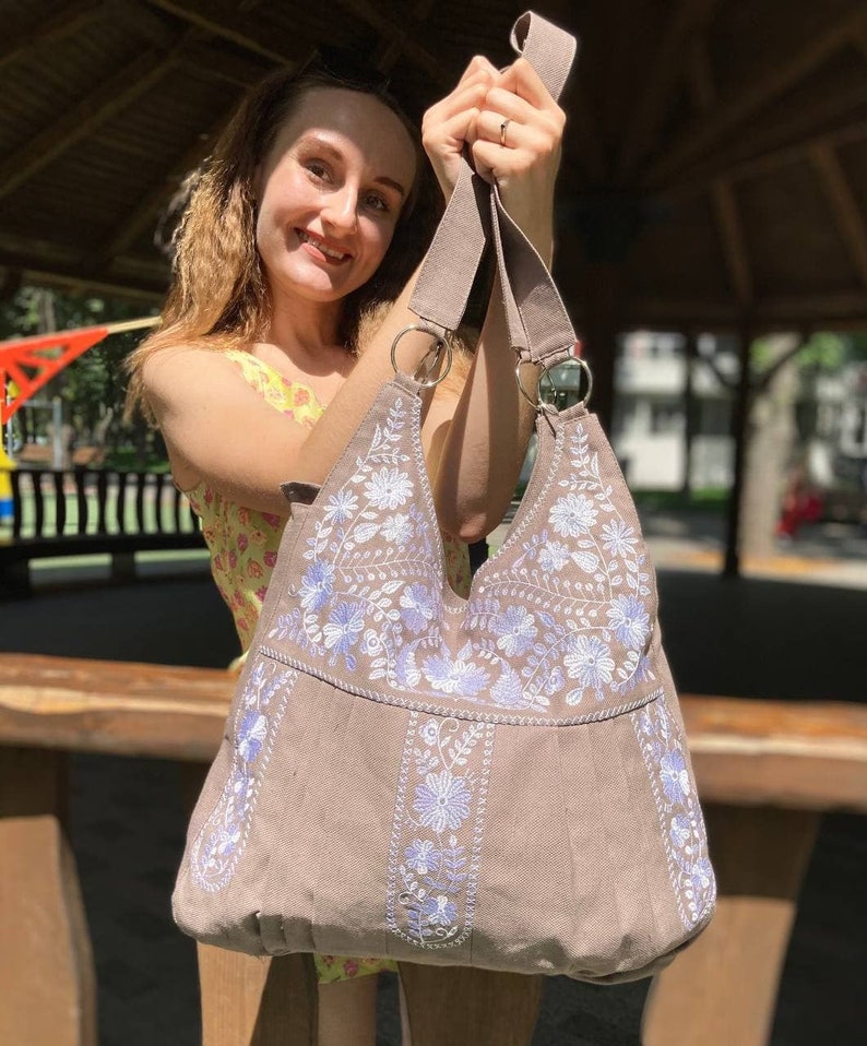 K&ouml;nnte beinhalten: Eine taupefarbene Umh&auml;ngetasche mit wei&szlig;en und hellvioletten Blumenstickereien. Die Tasche hat einen breiten Riemen und Metallringe. Die Tasche wird von einer Person gehalten.