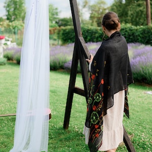 May include: A woman wearing a black shawl with red and green floral embroidery, standing near a wooden arch draped with white fabric. The scene is set outdoors with a lavender field in the background.