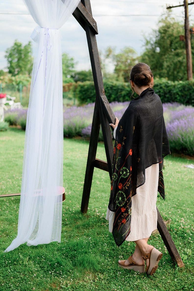 May include: A woman wearing a black shawl with red and green floral embroidery, standing near a wooden arch draped with white fabric. The scene is set outdoors with a lavender field in the background.