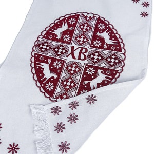 May include: White rectangular textile with a burgundy circular design featuring deer, snowflakes, and geometric patterns. The design includes the letters "XB". The edges are fringed, and small burgundy snowflakes are scattered around the border.