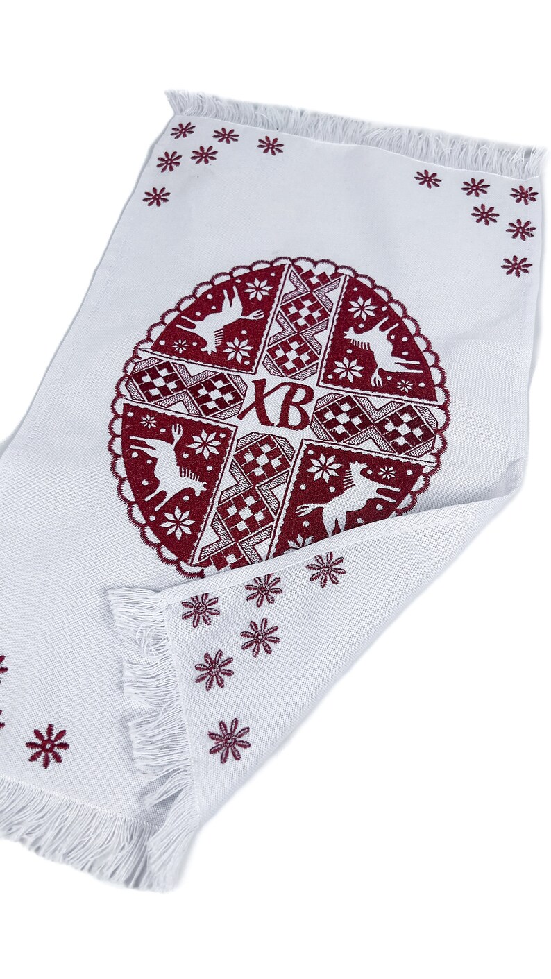 May include: White rectangular textile with a burgundy circular design featuring deer, snowflakes, and geometric patterns. The design includes the letters "XB". The edges are fringed, and small burgundy snowflakes are scattered around the border.