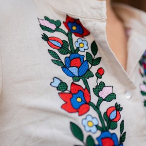 May include: Cream-colored shirt with colorful floral embroidery. The embroidery features red, blue, and pink flowers with green leaves. The shirt has a button-down front and a collar.